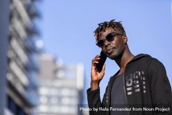 Serious Black man with sunglasses standing against cityscape on the street while using a mobile phone in sunny day bDjaX8