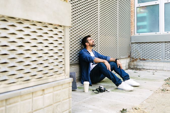 Latino man in denim sitting on ground outside with cup of coffee