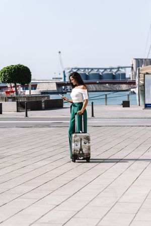 Side view of stylish young woman on the mobile phone with suitcase standing on the street