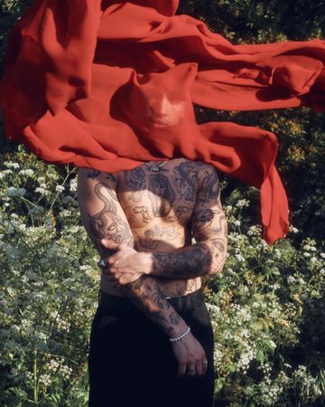 Tattooed male standing in garden with red sheet hiding his face, vertical