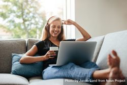 Smiling young woman enjoying a cup of coffee while relaxing with her laptop in living room 5R1QN0