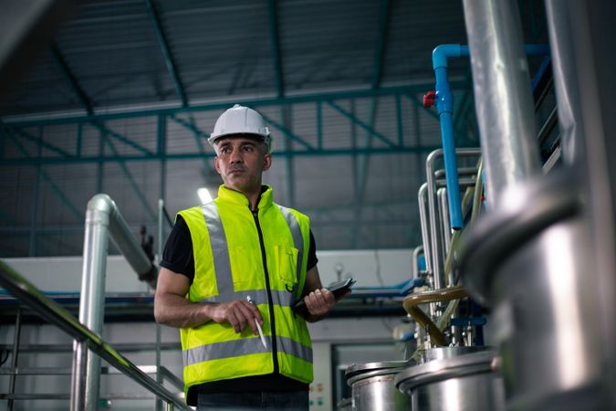 A male industrial engineer wearing a safety helmet and reflective vest is inspecting equipment in a factory. He is using a digital tablet to review data. Background show industrial pipes and machinery