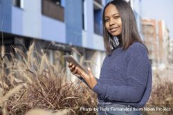 Confident female in sweater standing outside blue building in long grass with tablet 5zWmg4