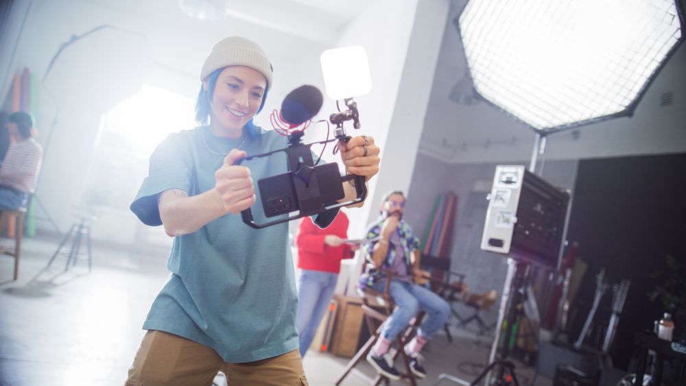 Young Female Videographer With Blue Hair, Wearing A Beanie, Captures Content In A Studio, Surrounded By Professional Lighting Equipment And Team Members Engaged In A Creative Production.