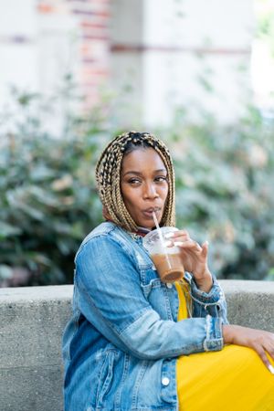 Portrait of woman drinking iced coffee with a straw sitting on stone bench outside
