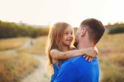Man carrying daughter in long grass meadow at dusk