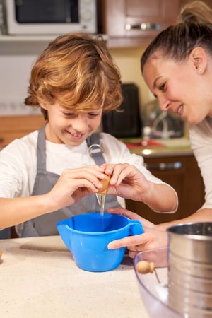 Happy boy breaking egg into container while cooking with mom