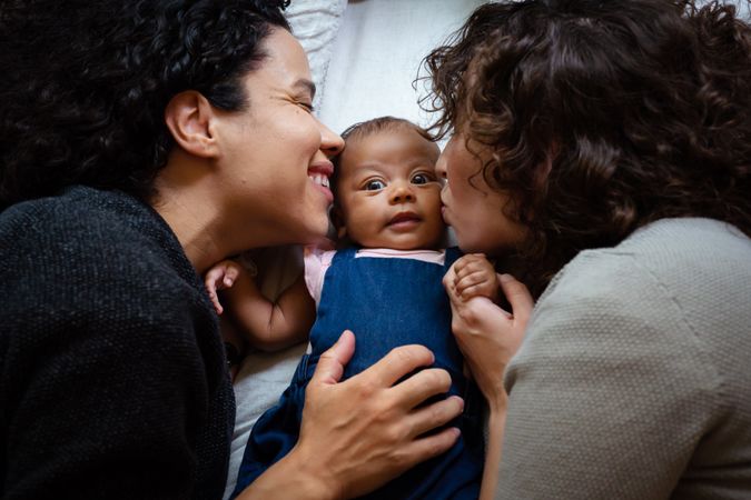 Close up of two moms kissing a baby girl while lying on the bed