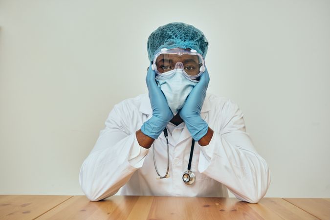 Tired Black male doctor in ppe gear at a table resting his head in his hands looking at camera