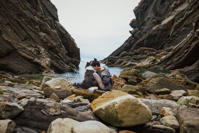 Traveling woman kissing dog sitting on boulder coast