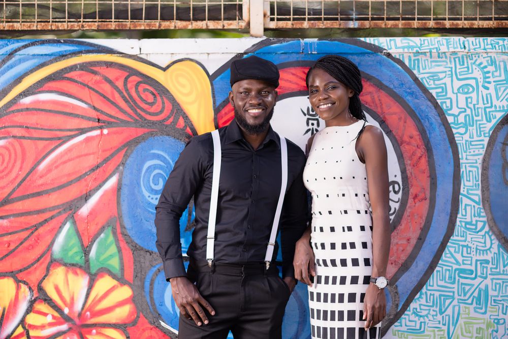 Man in dark shirt with strap and hat and a woman in light dress smiling