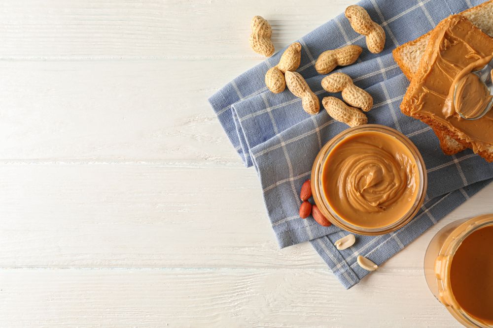 Glass jar with peanut butter, peanut, kitchen towel, spoon and peanut butter sandwich on wooden background, top view and space for text