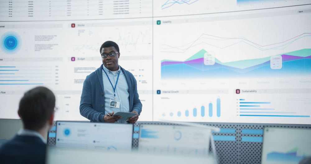 Black Male Financial Analyst Giving Presentation To Venture Capital Company Employees On Big Digital Screen In Monitoring Room.