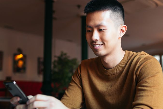 Smiling Asian man sitting in restaurant looking at smartphone.