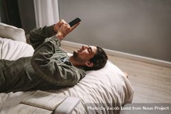 Young man using mobile phone lying on bed with books by his side bEnBG0