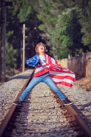 Joyful preteen boy wrapped in American flag standing on rails