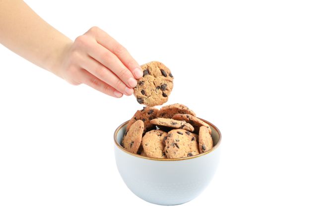 Young woman holding tasty chocolate chip cookie over bowl on plain background, closeup
