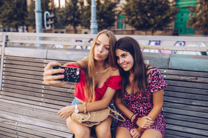 Laughing women taking selfie on wooden bench at street