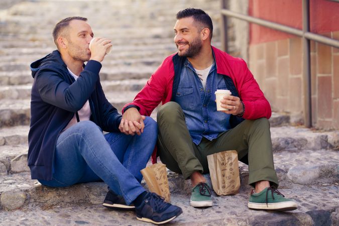 Two men holding hands and sitting outside with to go coffee