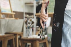 A close-up of the artist hands wearing an apron smeared with paint. Clutching many brushes and paintbrushes.