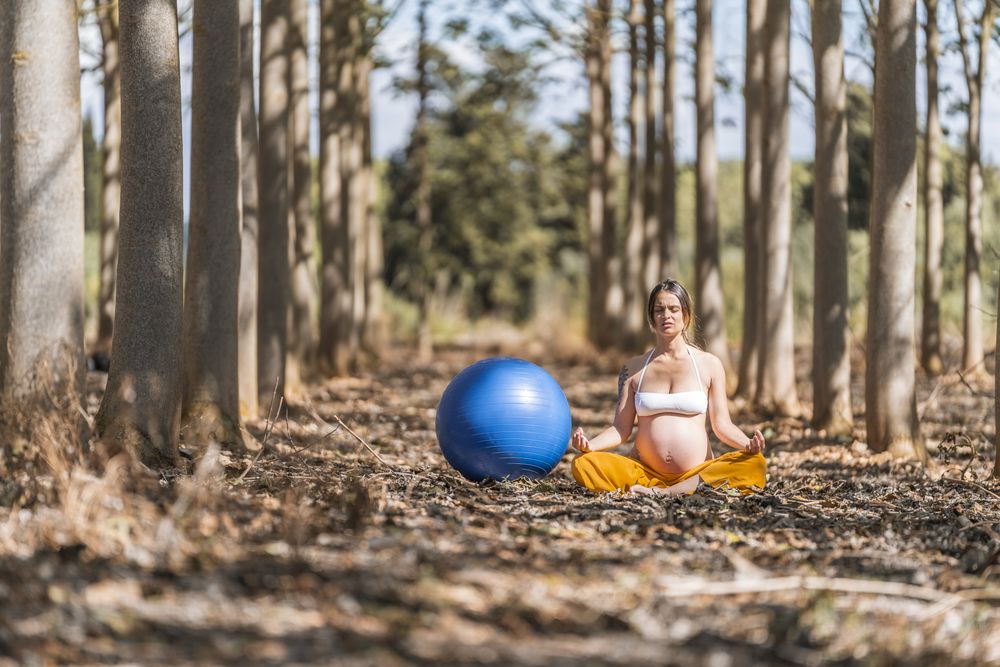 Serene adult pregnant woman meditating while sitting on ground among trees in park during sunny day