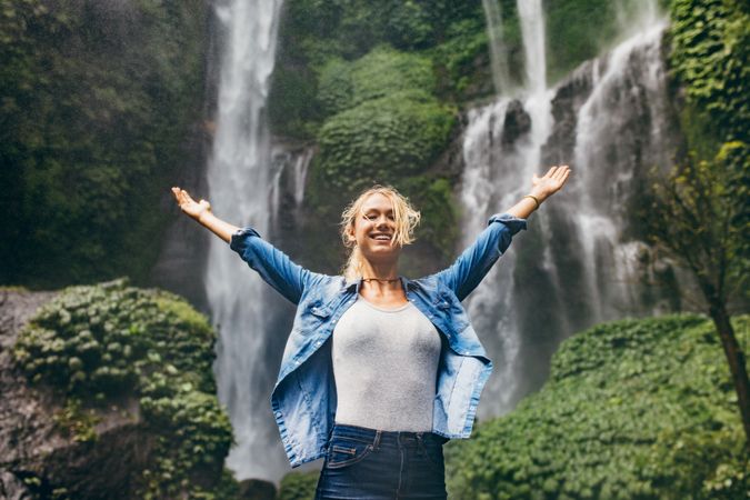 Happy young woman spreading hands enjoying nature with waterfall in background