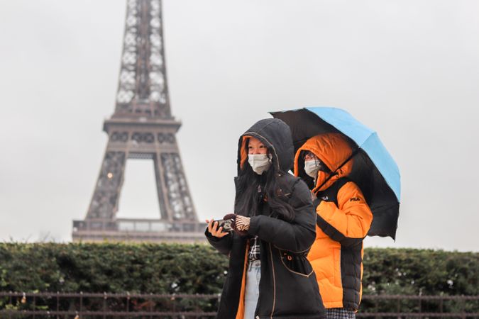 Two people with facemasks wearing jackets walking under the rain near Eifel tower in Paris, France