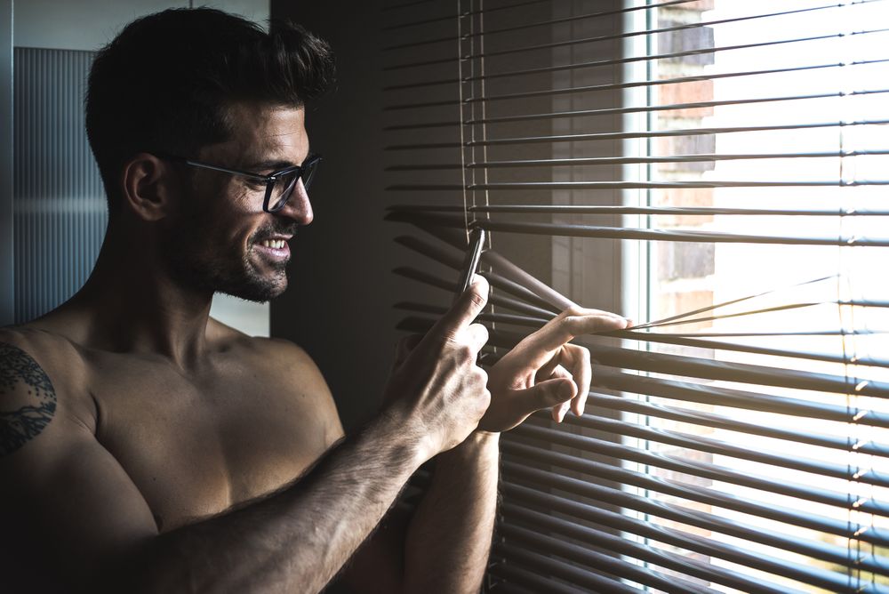Man standing by a windows blind on the mobile phone