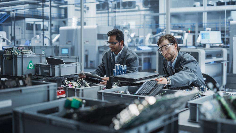 Indian And White Male Workers Taking Apart Laptops To Recycle For Production Of High Tech Devices At Automated Electronics Factory With Robotic Arms.