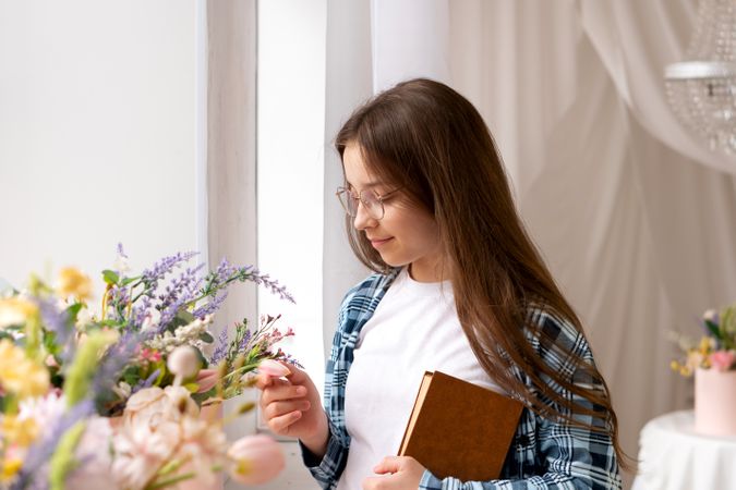Teen girl holding book touches flowers near bright window.