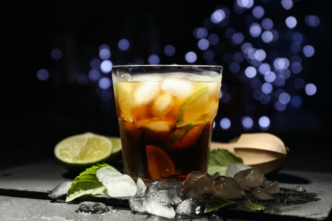 Glass with cola and lime slices, juicer, ice, mint against blurred lights background