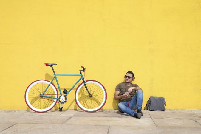 Male looking around in front of yellow wall next to bike with bike and listening to music