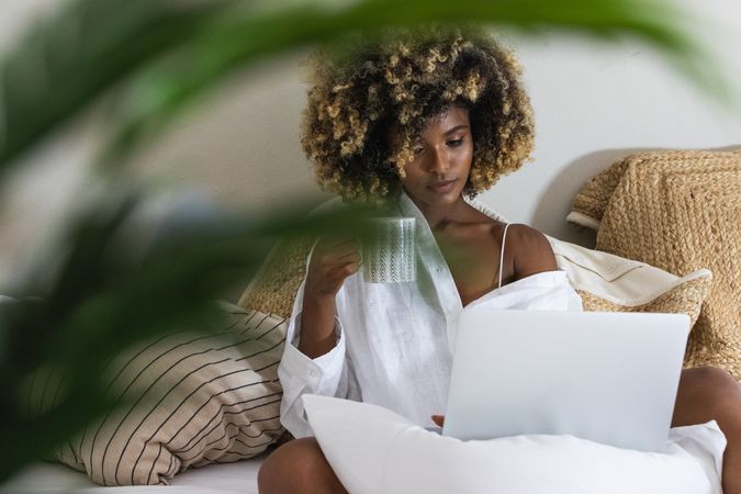 Black woman drinking coffee while sitting on bed with laptop