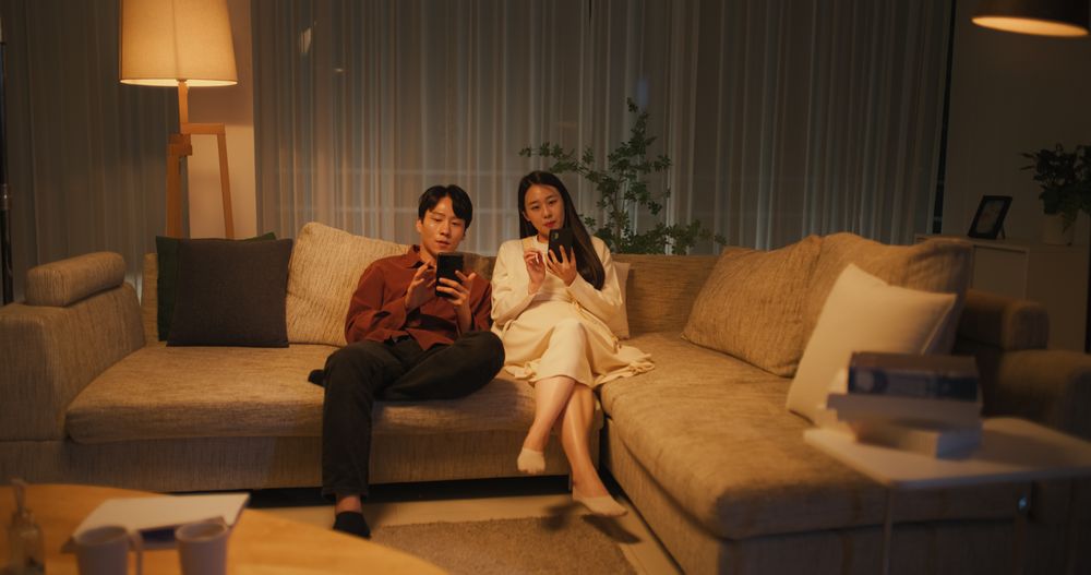 South Korean Couple Enjoying Leisure Time at Home in the Evening, Both Using Smartphones For Online Shopping