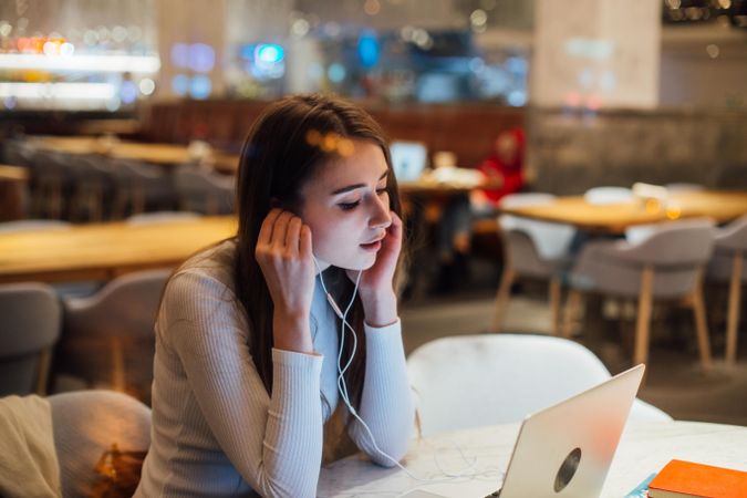 Contemplative brunette woman in a restaurant working on her laptop with earbuds