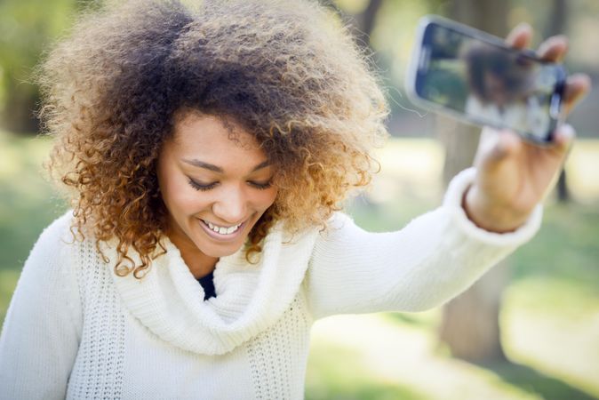 Smiling Black woman taking a selfie on her phone in bright park while looking down