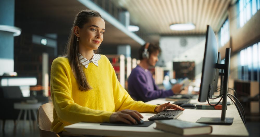 Female Student in Yellow Jumper Working on Her School Thesis on a Desktop Computer. Young Smart Woman Studying Online in a Quiet Public Library with Book Shelves