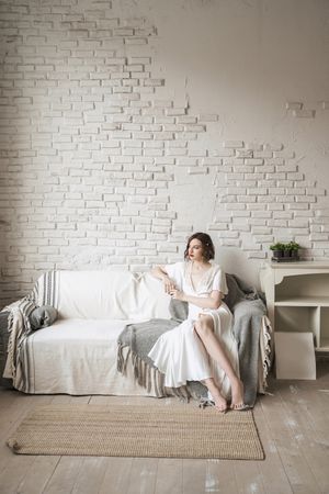 Pensive adult woman looking away and dreaming while sitting on sofa and having hot drink in light modern living room