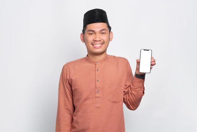 Muslim man in kufi hat standing tall holding mobile phone with mock up screen