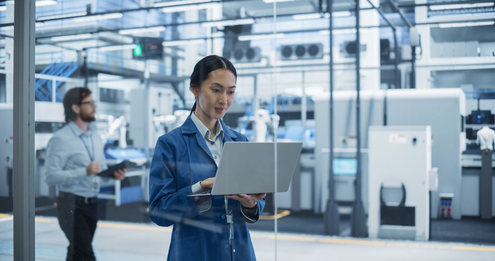 Electronics Manufacturing Technician in a Blue Coat Working on a Laptop Computer at an Assembly Plant. Asian Female Specialist Developing, Maintaining, Setting Up Software for Automated Production