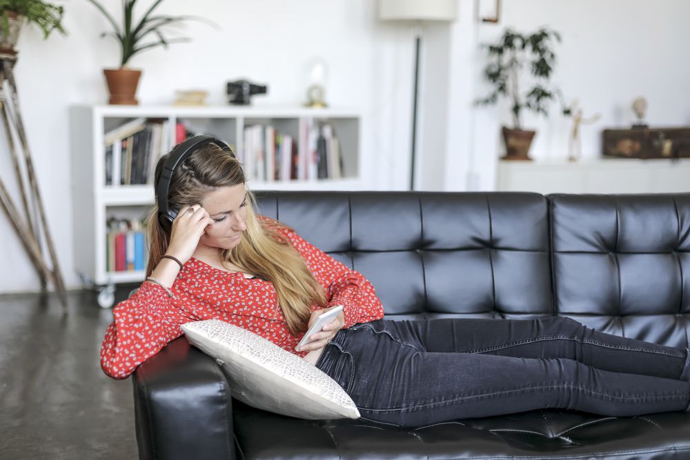 Blonde woman using mobile and listening to music by headphones while lying on leather sofa