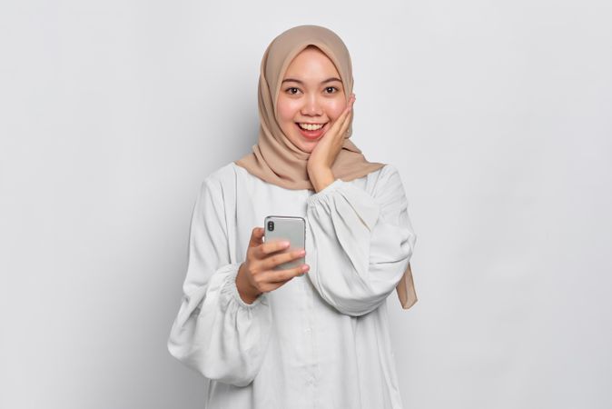 Asian female in headscarf receiving happy news on her cell phone with hand on her cheek