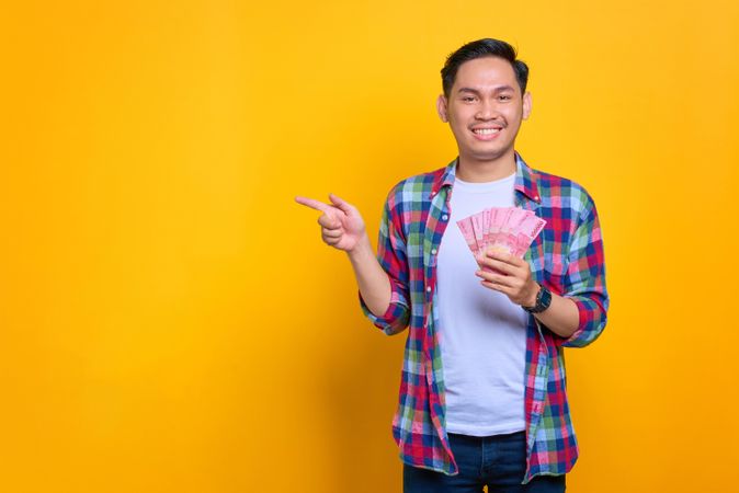Smiling Asian man holding up cash and pointing to copy space in studio shoot