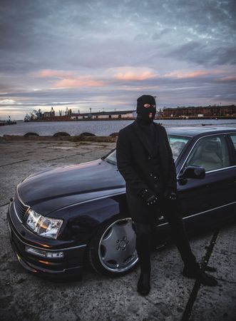 Man in dark suit and robber mask leaning on a dark car