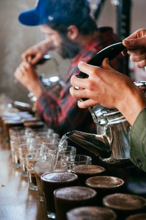Hands pouring hot water for multiple coffee tastings with man blurred in background