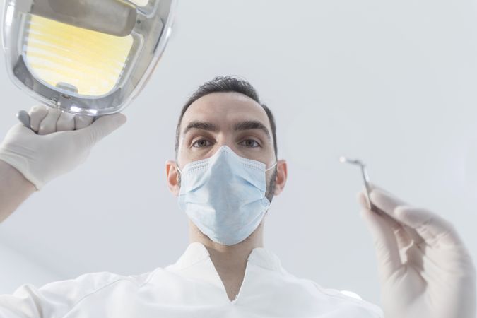 Dentist wearing surgical mask while holding angled mirror and drill, ready to begin work