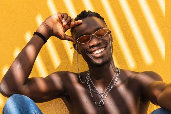 Cheerful Black man in trendy sunglasses sitting near wall