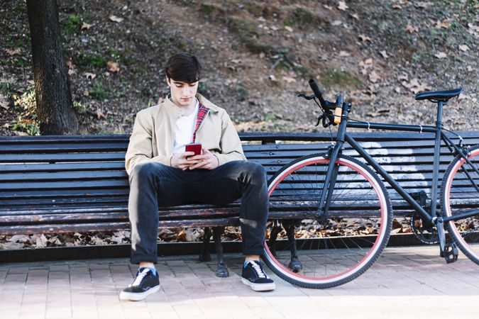 Man sitting on bench outside next to bike checking phone