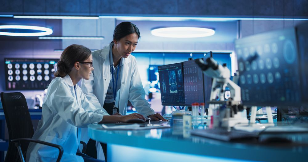 Asian And White Female Doctors Talking In Medical Research Center And Using Desktop Computer To Analyze MRI Scans Of Brain.