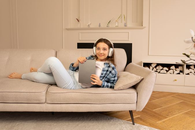 Teenage girl lounging on sofa wearing headphones holding tablet device.